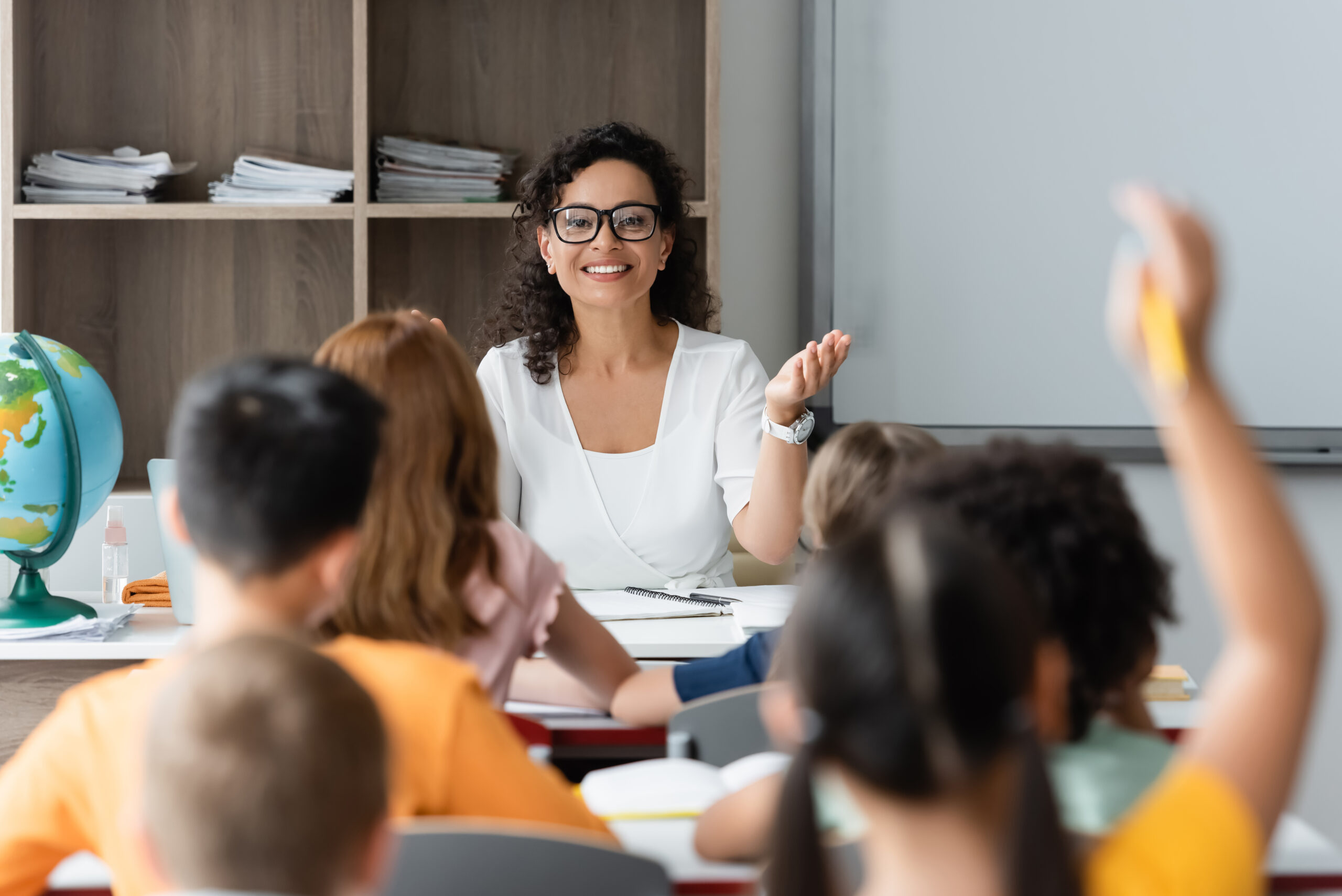 positive teacher pointing at blurred girl raising hand in classroom