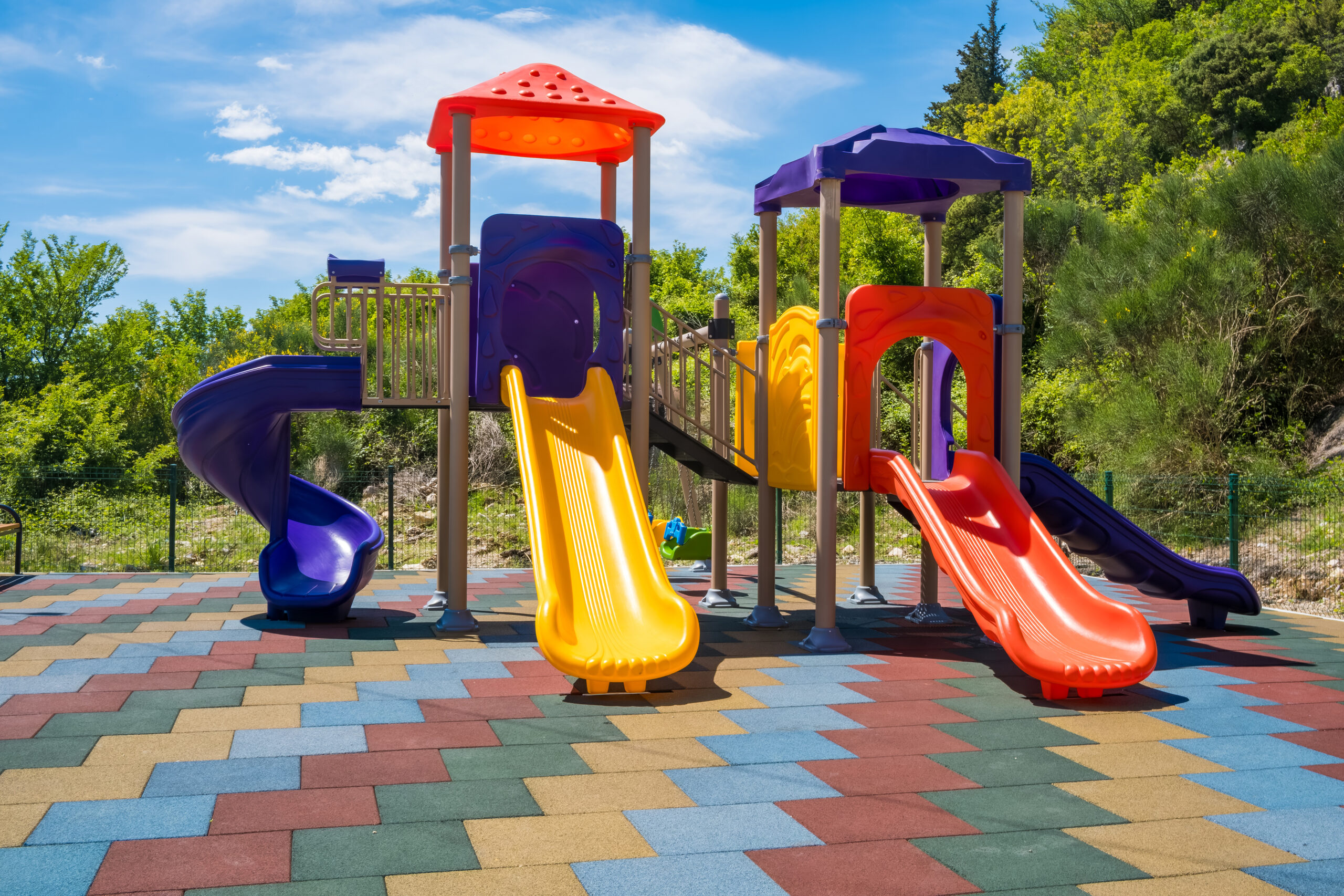 Colorful playground with slides and climbing structures under a blue sky. The ground is covered with multicolored rubber tiles.
