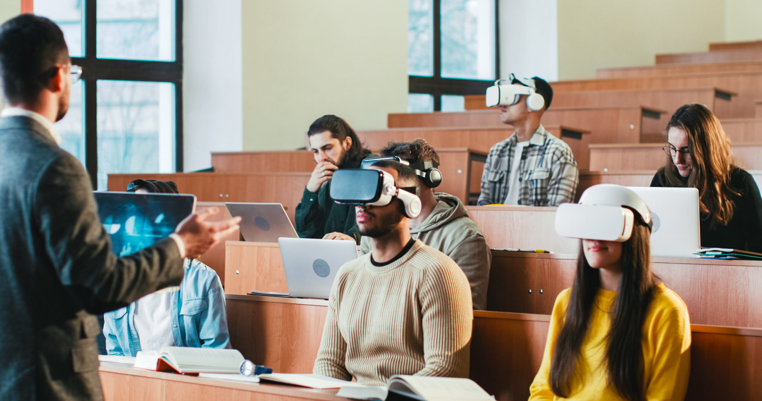 Rear of professor having speech in front of students in VR glasses with laptops.